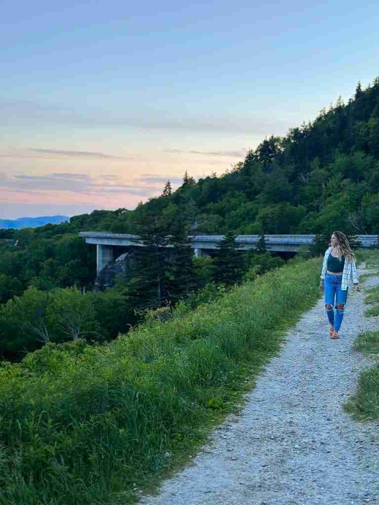 Girl walking near the Blue Ridge Parkway with the Linn Cove viaduct (a bridge) in the background at sunset. 